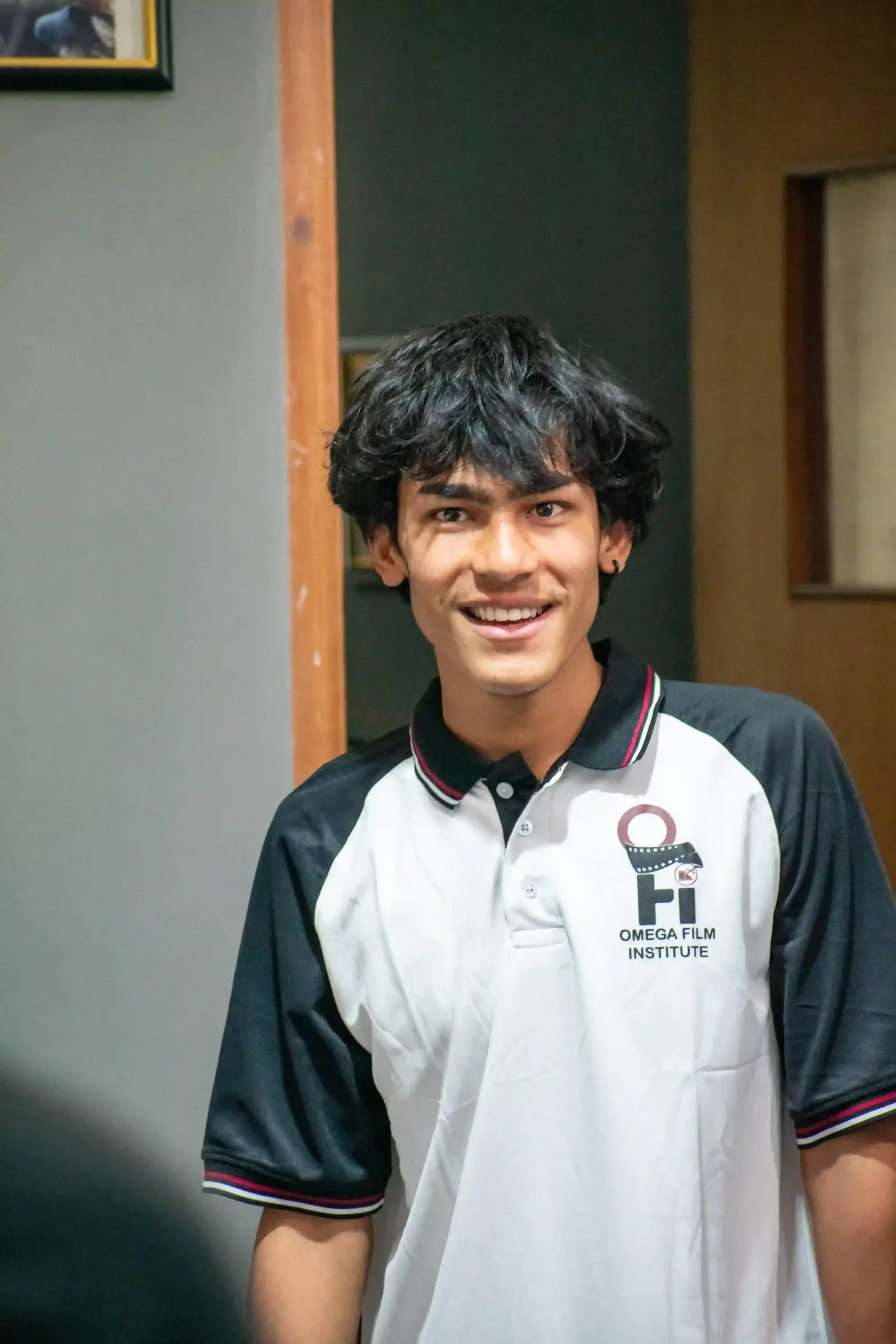 Young man with black hair smiling, wearing a white and black polo shirt with an Omega Film Institute logo, standing by a doorway.